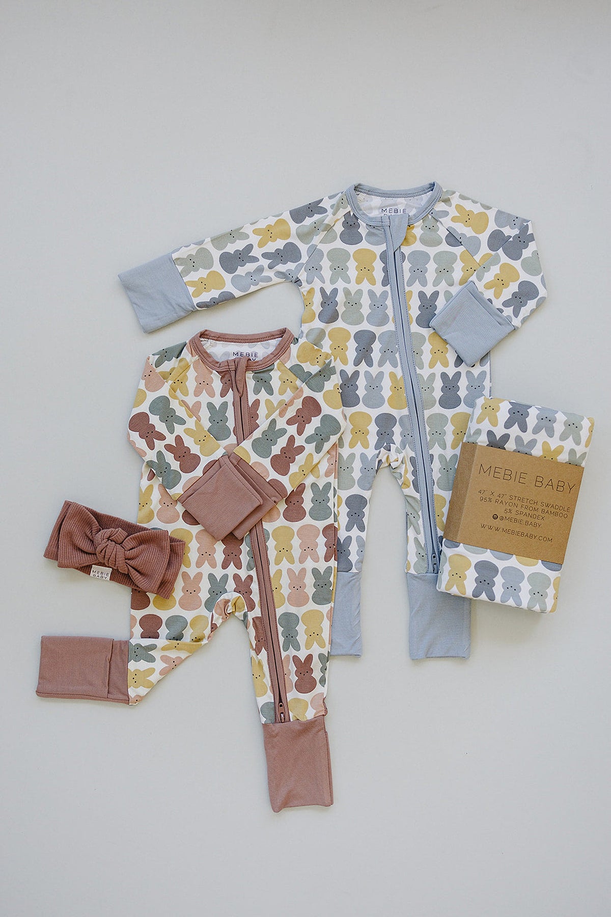 Two Mebie Baby zipper pajamas with colorful bunny patterns, one with dusty blue accents, paired with a matching headband.