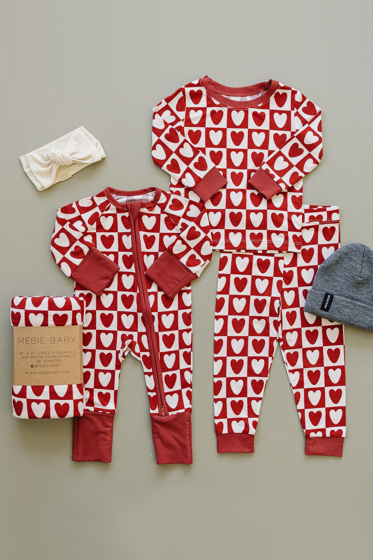 A set of Mebie Baby checkered heart ribbed pajamas in red and white, featuring a zipper sleeper and matching top and pants, arranged with a headband and blanket.
