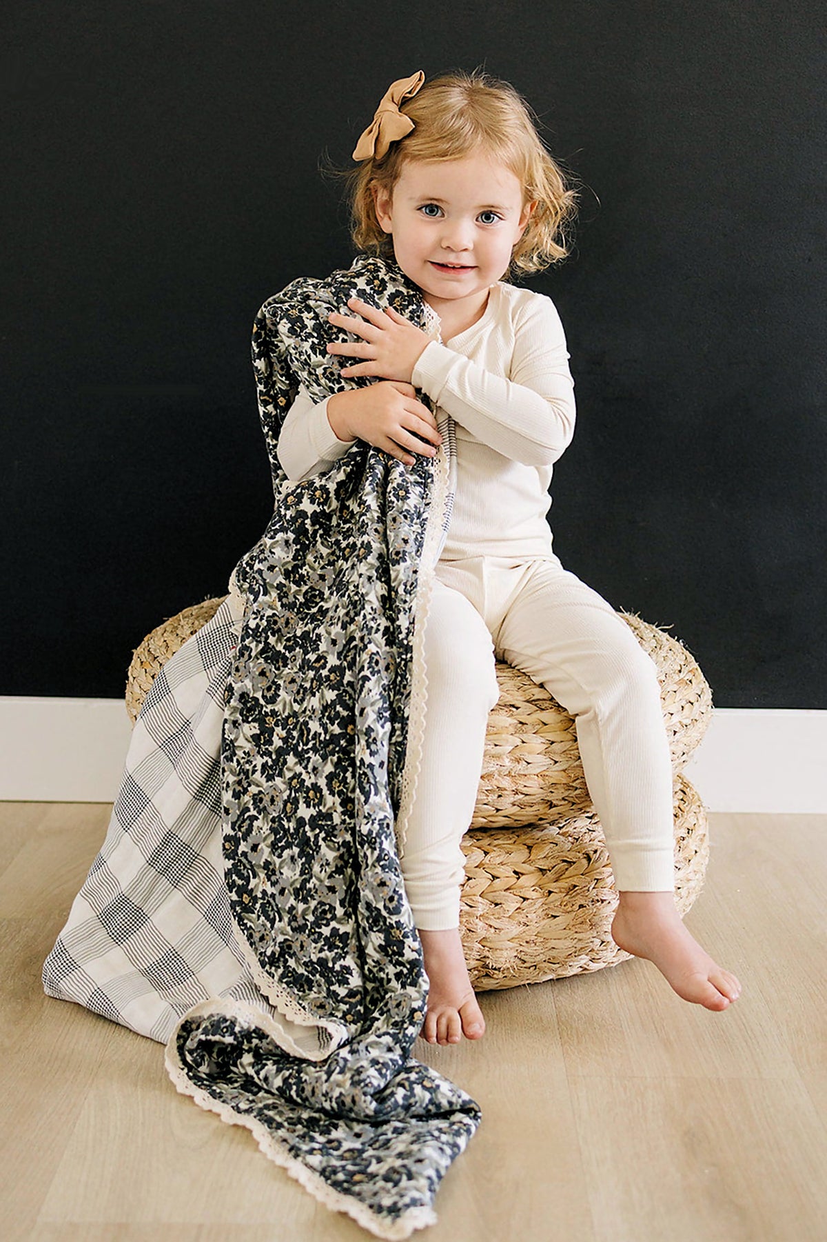 A child in cream pajamas sits on woven stools, holding a floral and plaid quilt with lace edges.