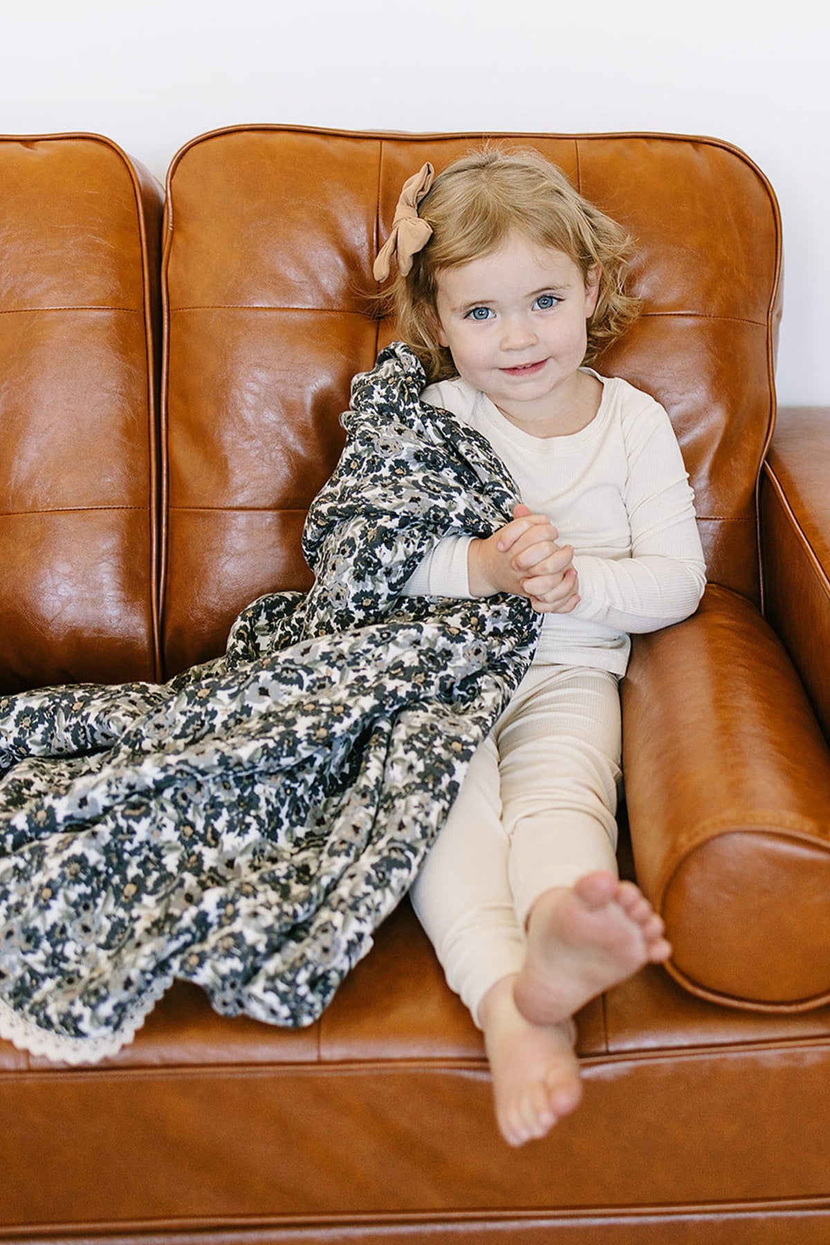 A child in light clothing sits on a brown leather couch, holding a floral-patterned blanket.