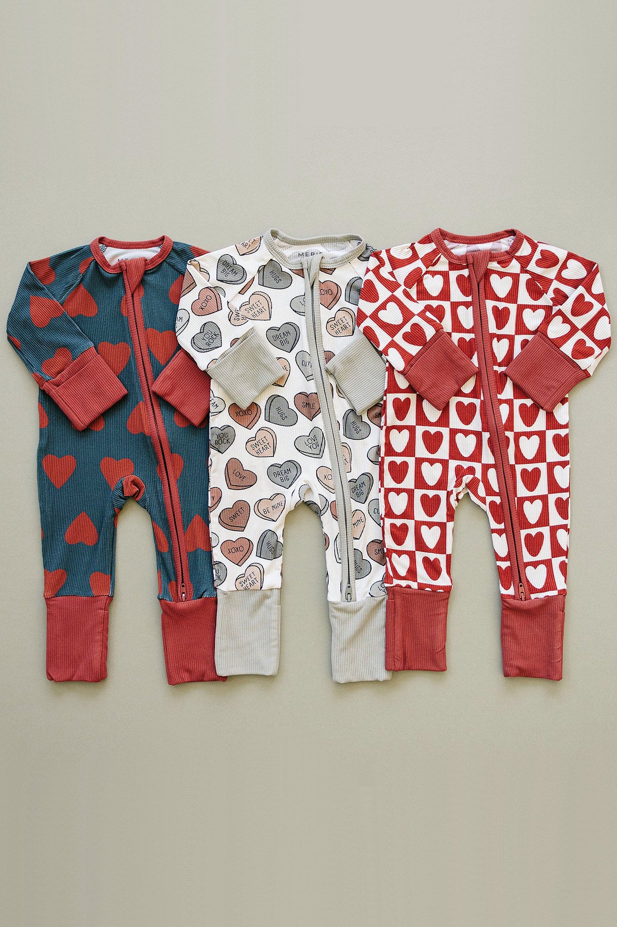 Three adorable baby zippered pajamas in varying heart patterns are displayed on a neutral background. The pajamas feature soft bamboo fabric, making them cozy and perfect for Valentines Day.