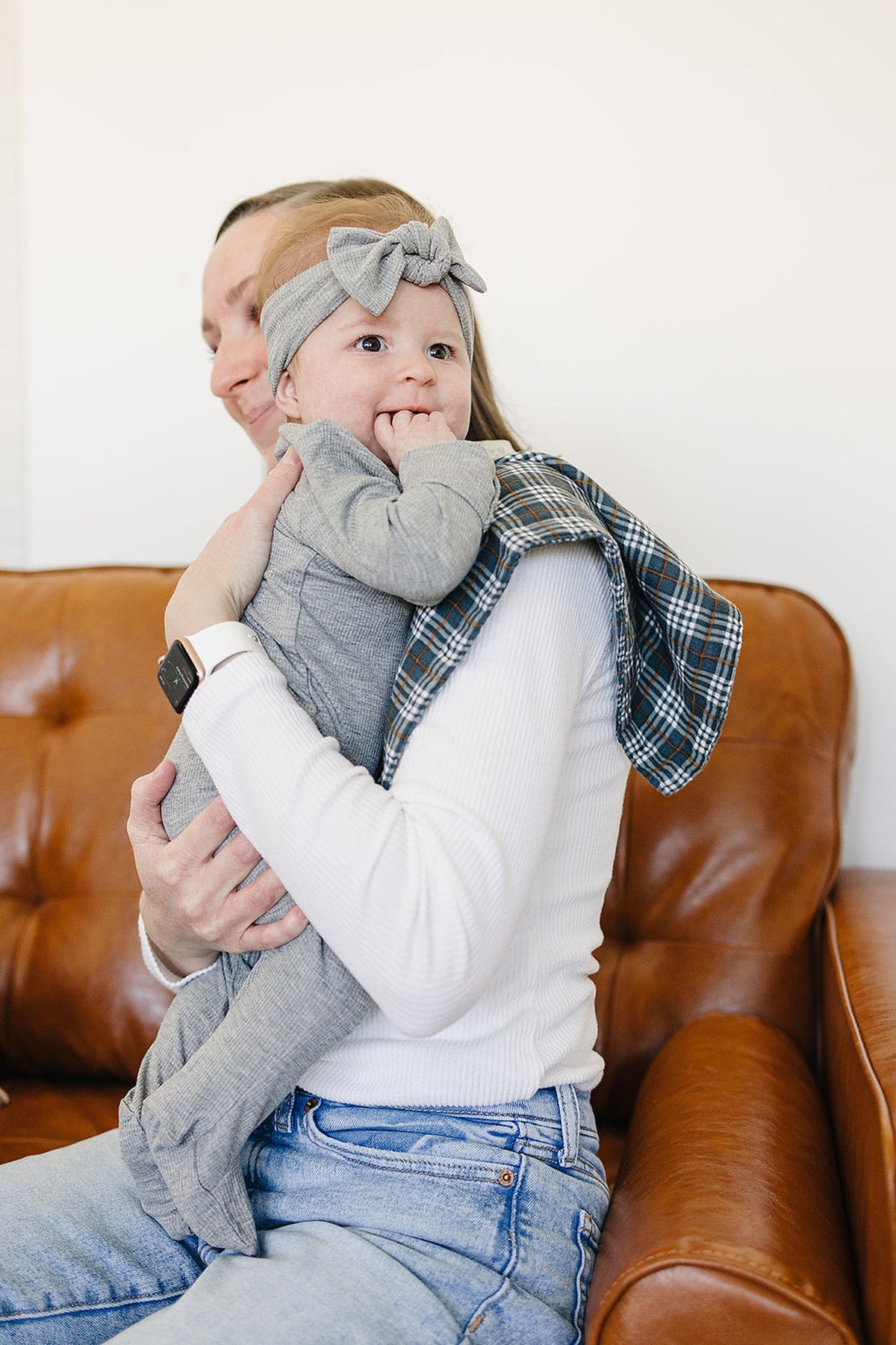 A parent holds a baby wearing a gray outfit and a matching headband, while a navy plaid muslin burp cloth drapes over the parents shoulder.