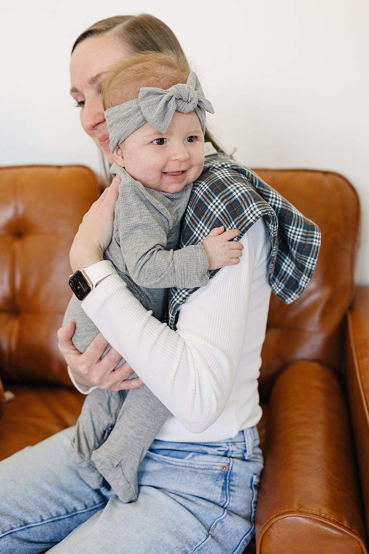 A smiling baby in a gray outfit and headband is held by a woman in a white top, sitting on a brown leather couch.