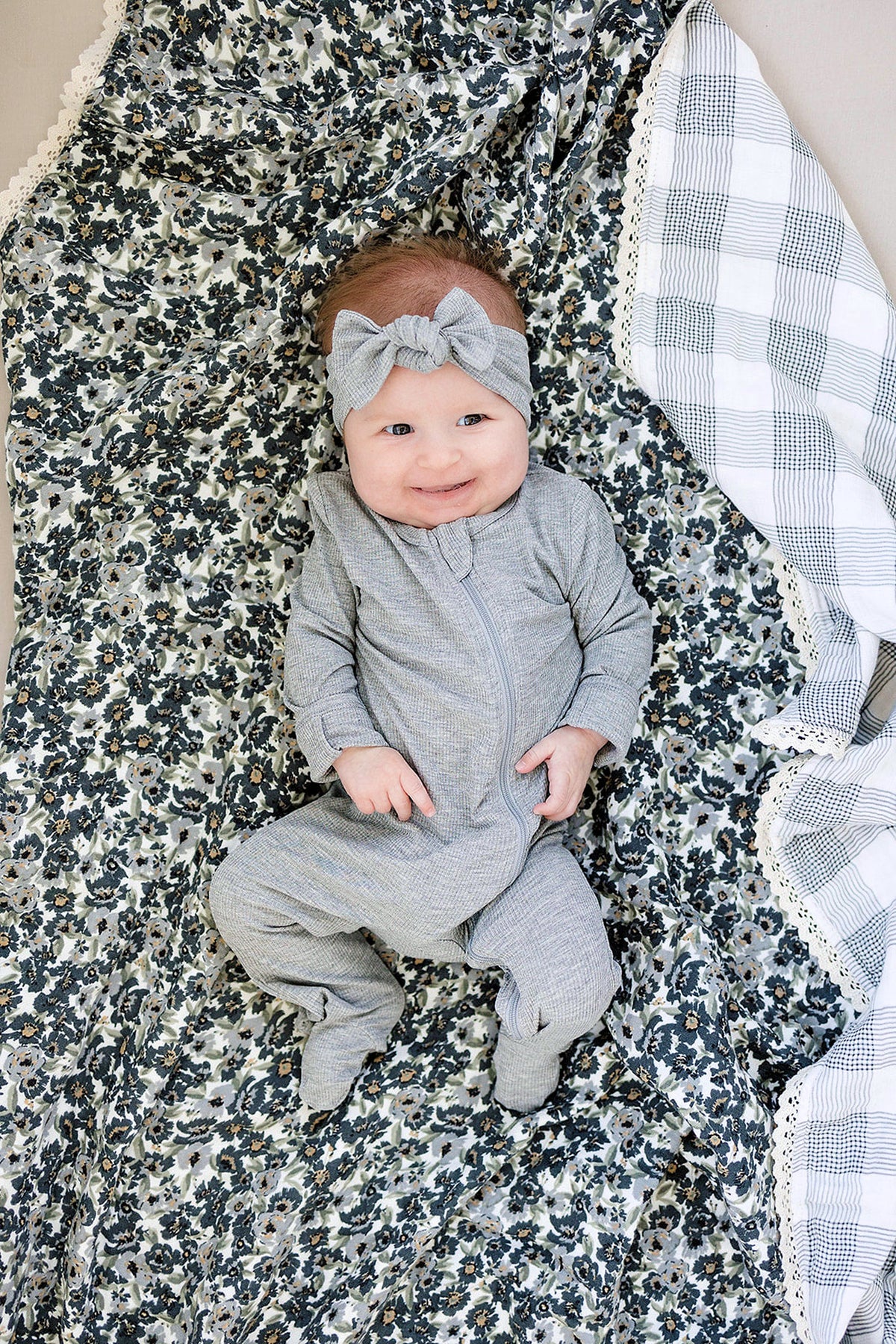 A baby in a gray outfit and headband lies on a floral and plaid quilt with lace edges.
