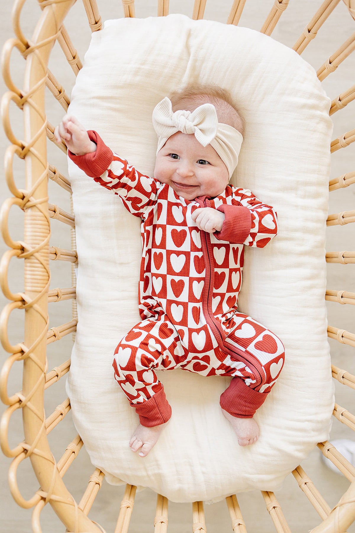 A smiling baby in a red and white checkered heart-patterned zipper pajama, made from soft bamboo fabric, is lying on a cream-colored cushion inside a woven bassinet.