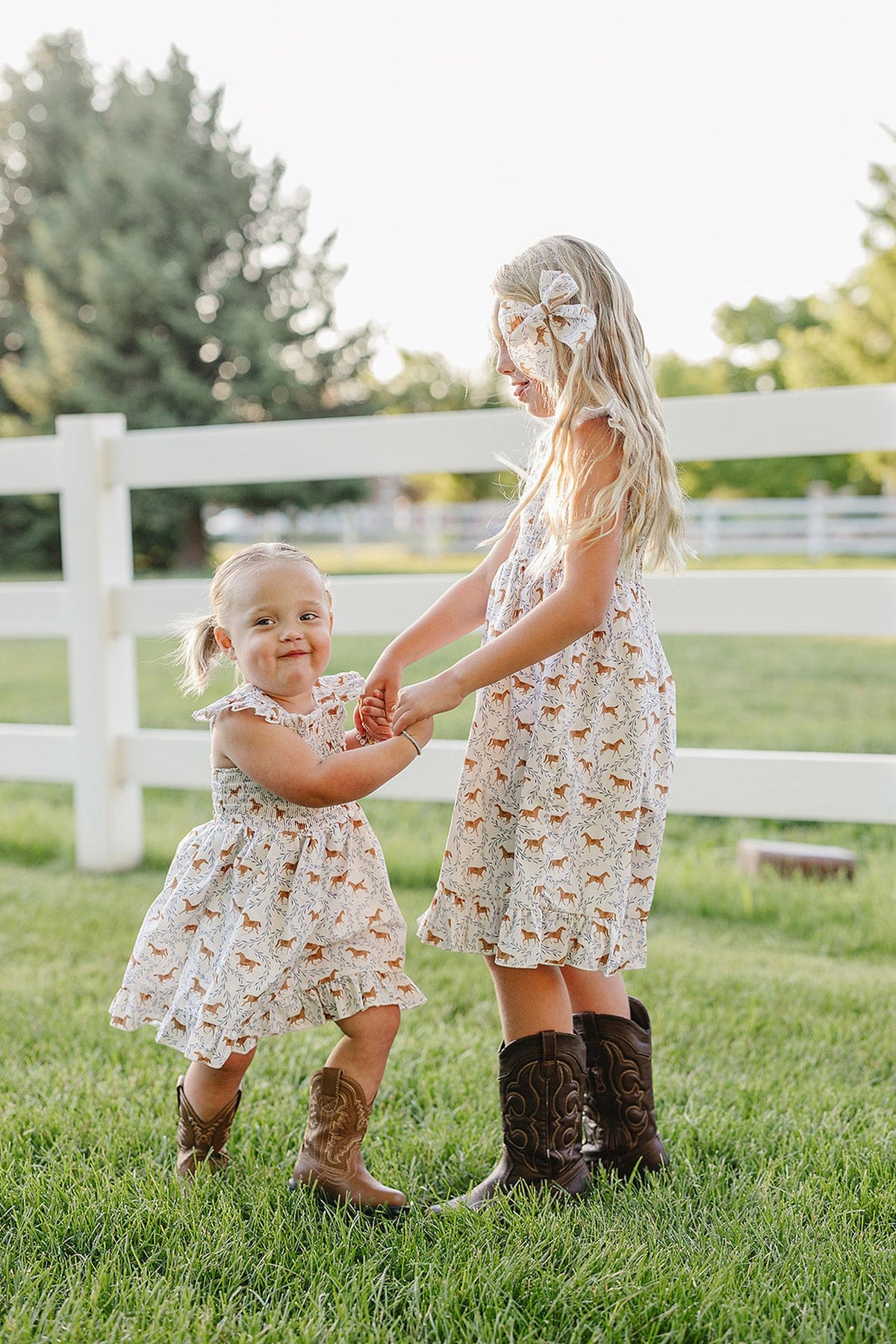 Two young girls are joyfully playing outdoors, wearing matching pony-printed smocked dresses, complemented by brown cowboy boots. The setting features a green lawn and a white fence in the background.