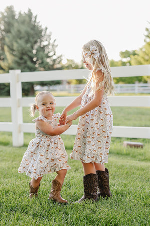 Two young girls are joyfully playing outdoors, wearing matching pony-printed smocked dresses, complemented by brown cowboy boots. The setting features a green lawn and a white fence in the background.