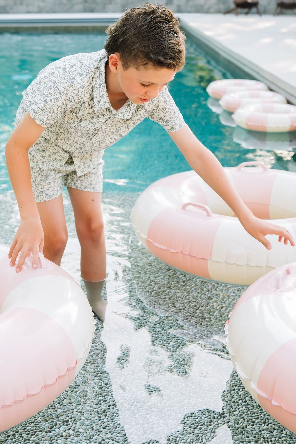 A child wearing a palm tree print shirt plays with pink and white pool floats in shallow water.