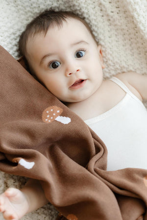 A baby lies on a cream knit blanket, wrapped in a brown swaddle featuring a mushroom design.