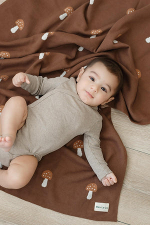 A baby lies on a brown mushroom-patterned blanket made from Pima cotton, wearing a beige onesie.