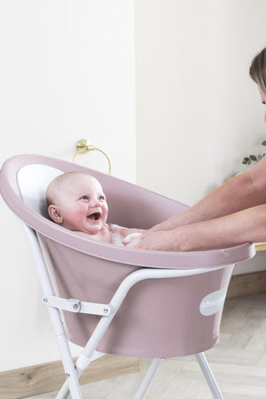 A smiling baby in a pink Shnuggle bath with a supportive backrest, being gently washed by an adult.