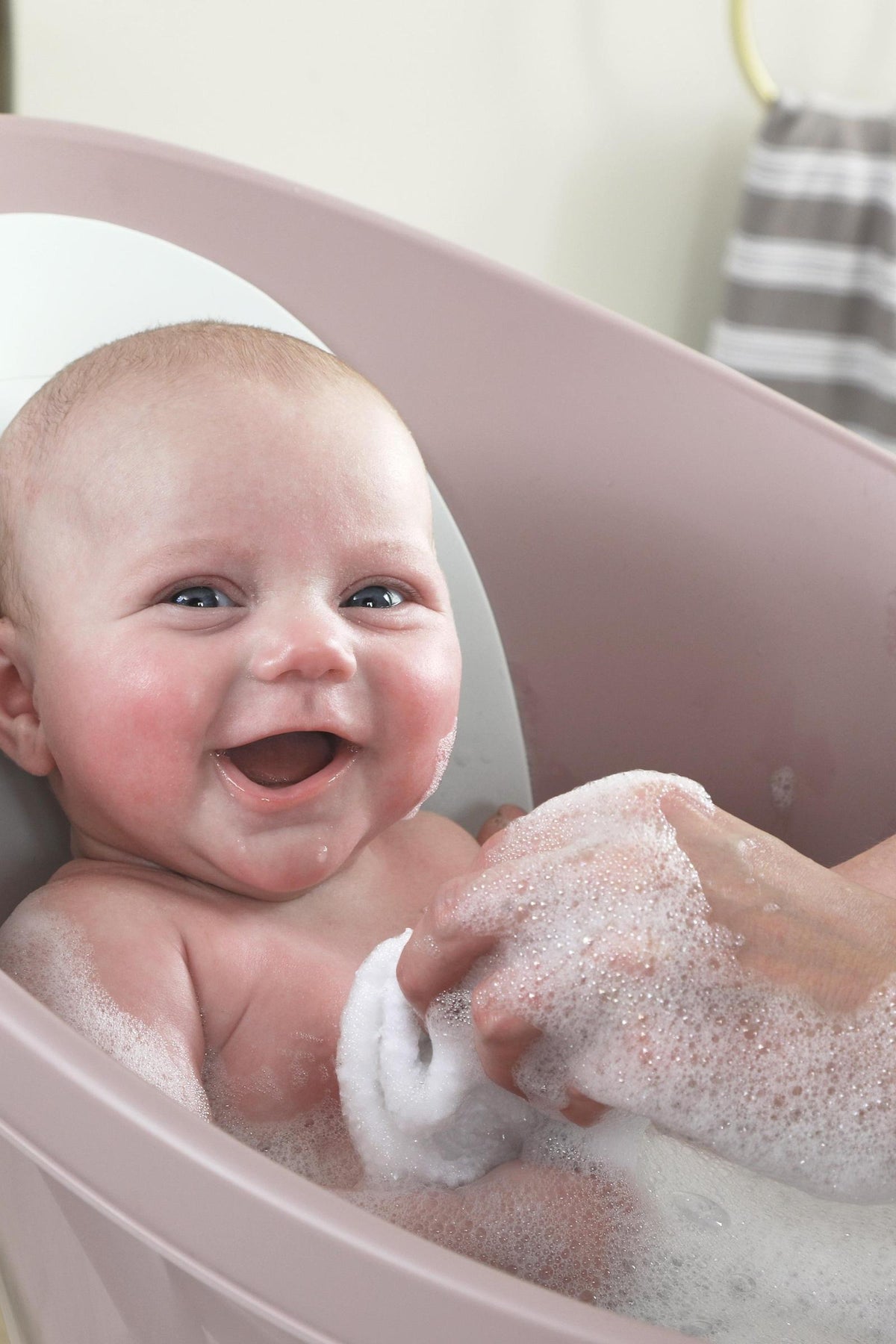 A smiling baby is bathed in a pink tub, with a hand gently washing them using a soapy cloth.