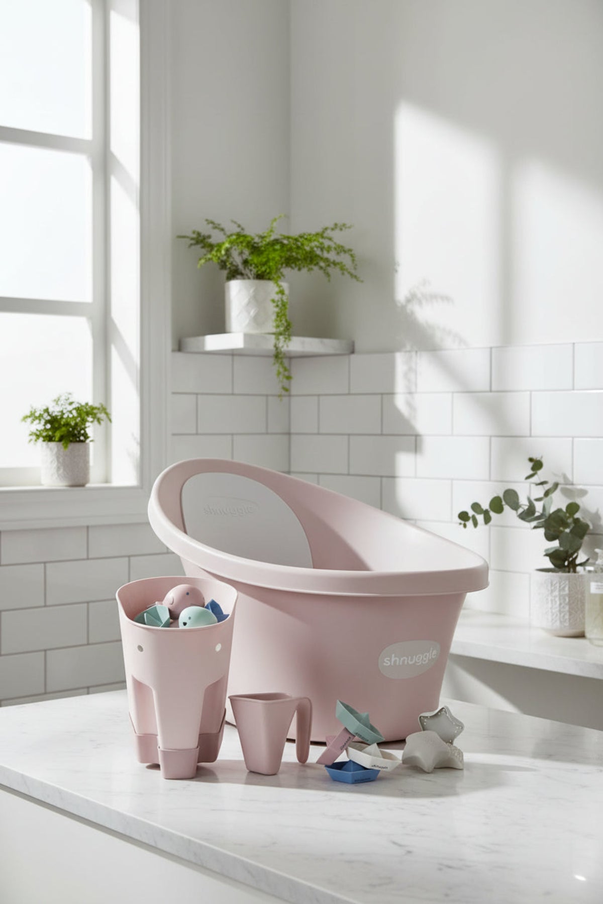 A pink Shnuggle baby bath set with toy accessories on a white countertop, surrounded by potted plants.