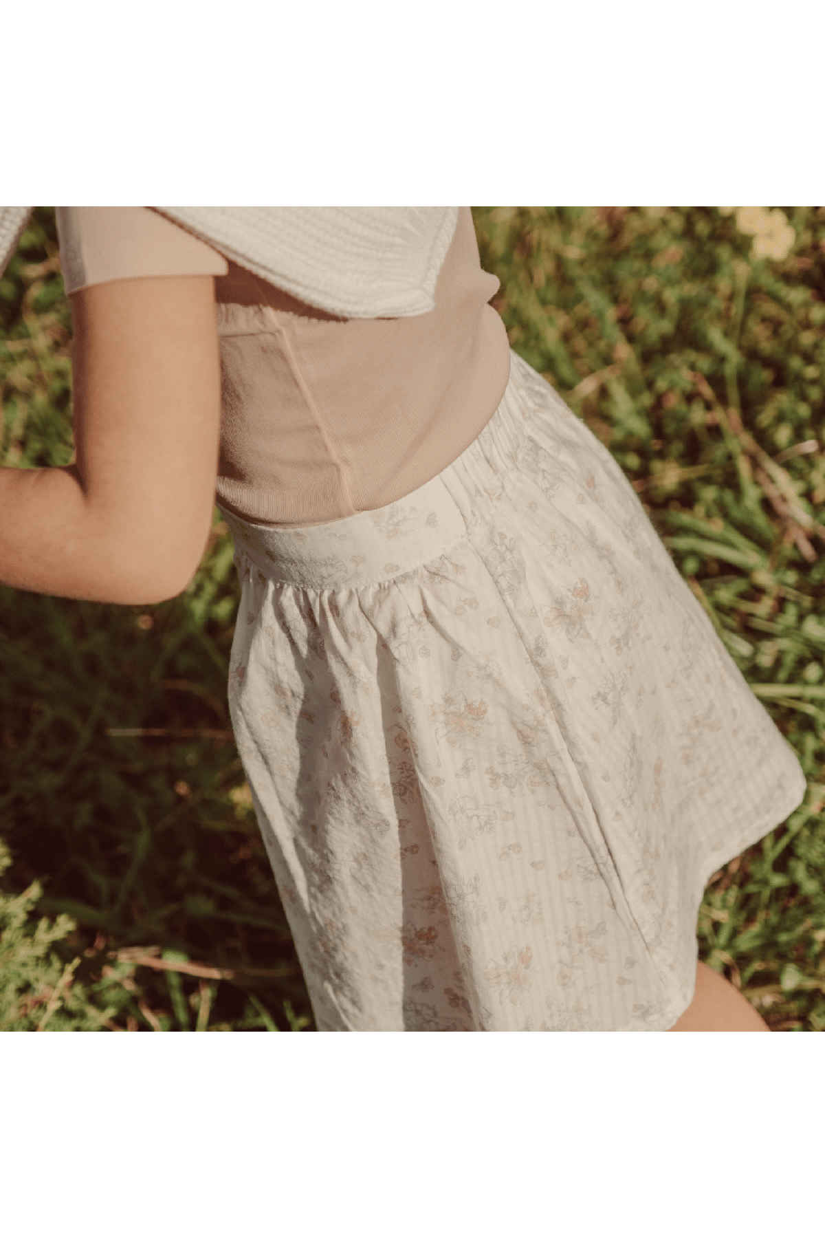 A young child wears a soft, floral printed skirt made from organic cotton, featuring a gathered design. The skirt is styled with a neutral top and surrounded by lush greenery.