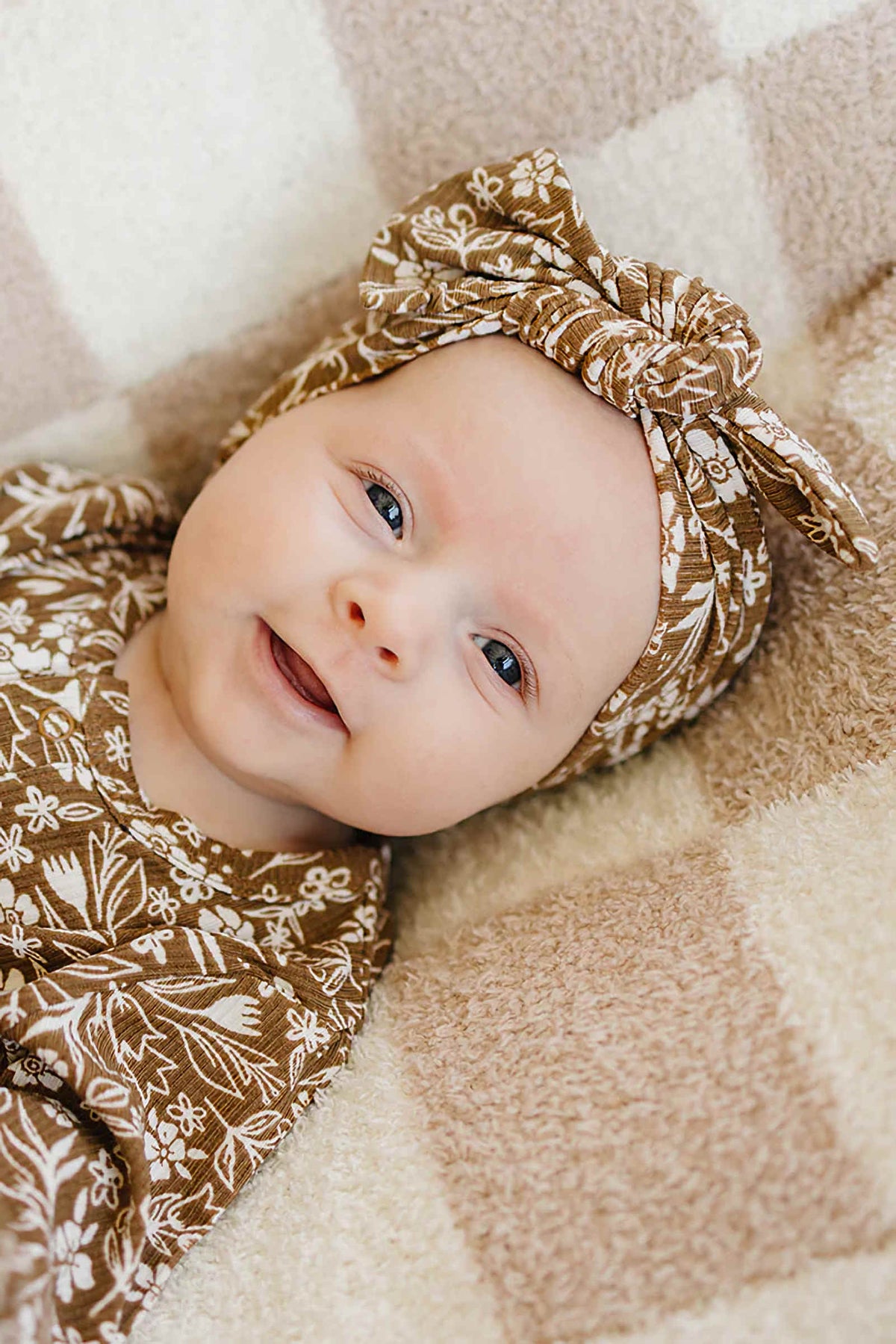 A smiling baby lies on a soft, beige checkered blanket, wearing a brown floral ribbed head wrap made from bamboo fabric, showcasing its gentle touch and stylish design.