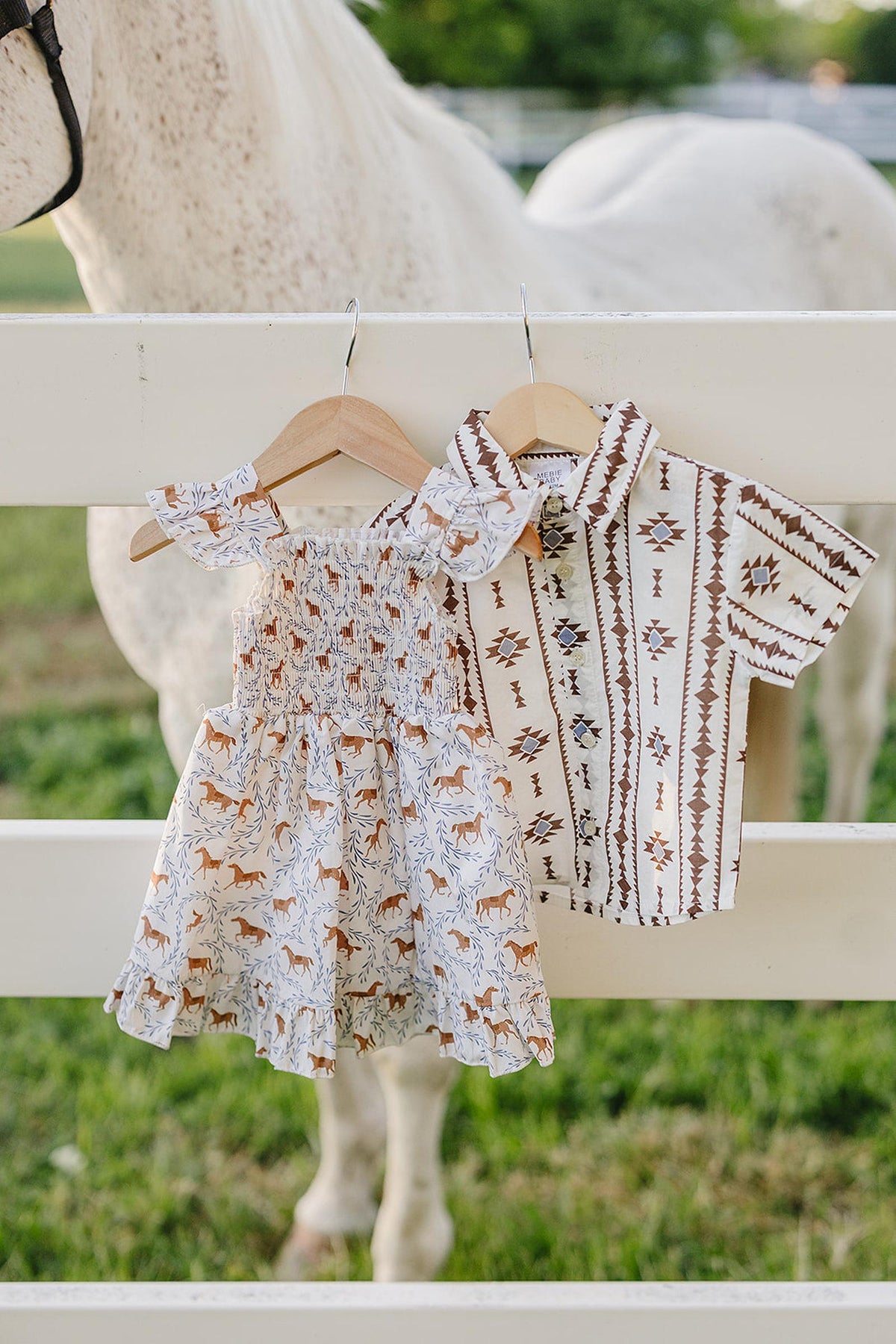 A smocked pony-print dress and matching shirt hang from wooden hangers against a white fence, with a horse in the background. The outfit features earthy tones suitable for young children.