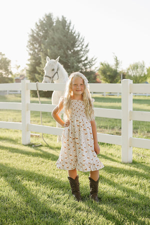 A young girl stands in a sunny field wearing a pony-printed smocked dress with ruffled hem and brown cowboy boots, beside a white horse and a white fence.