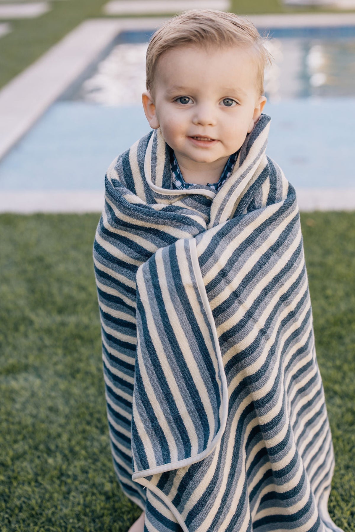 A child wrapped in a navy striped towel stands near a pool on a grassy area.