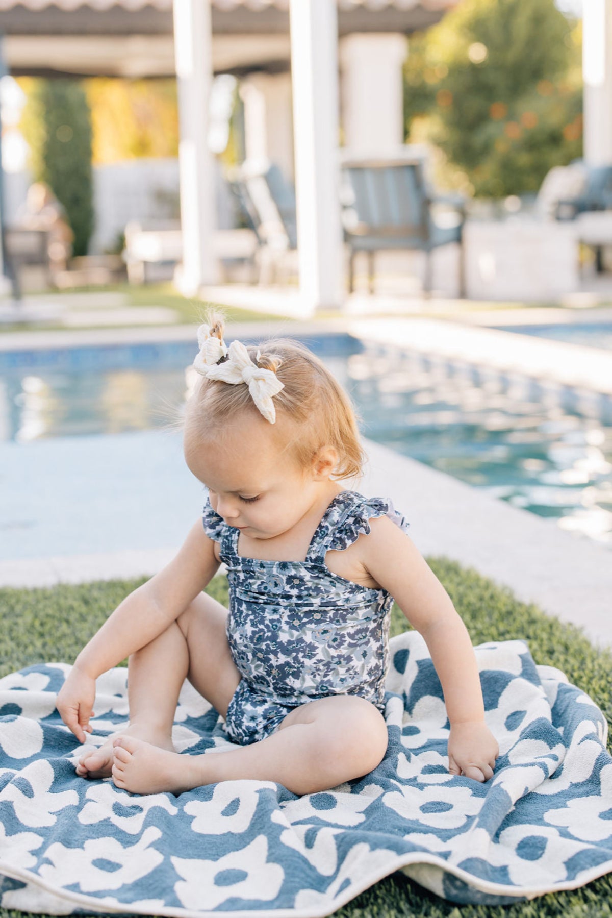 A toddler in a floral swimsuit sits on a navy floral towel by a pool, outdoors.
