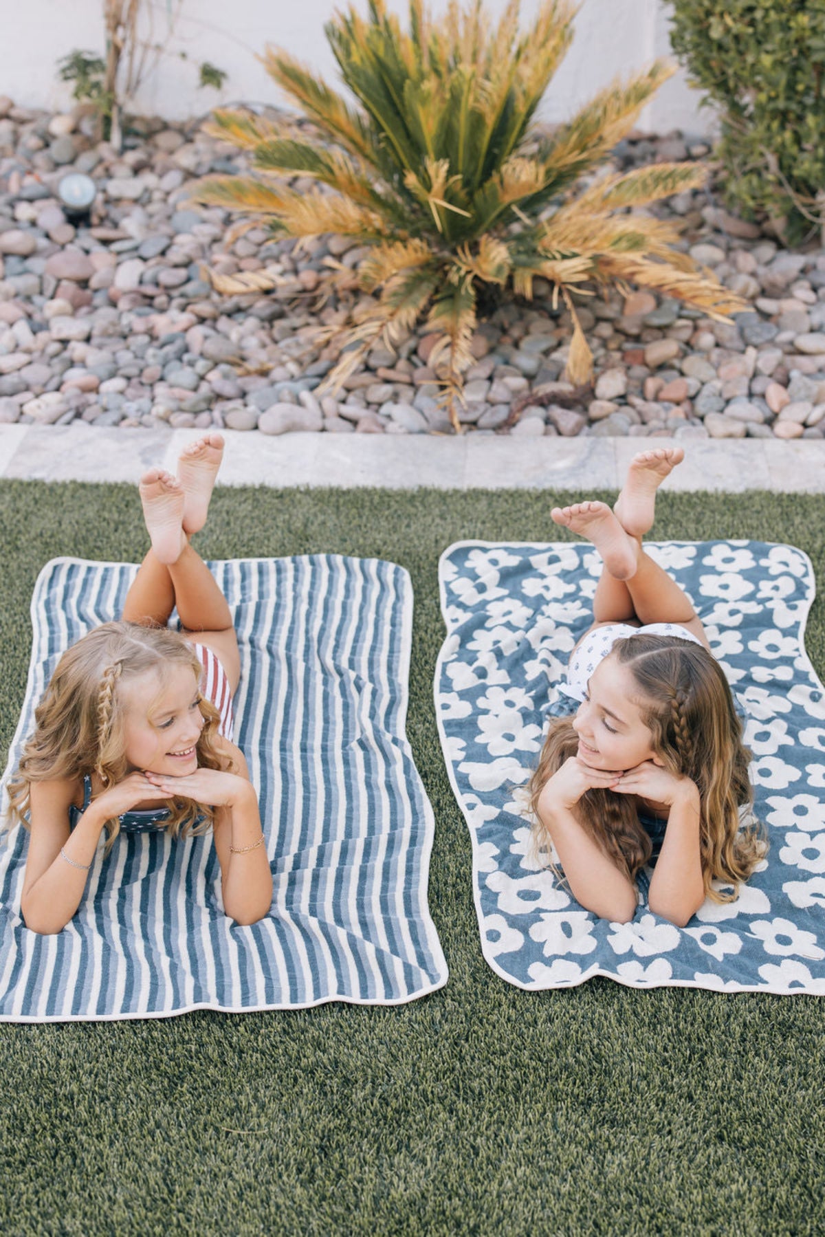 Two children lie on striped and floral towels on grass, smiling and relaxed near a garden.