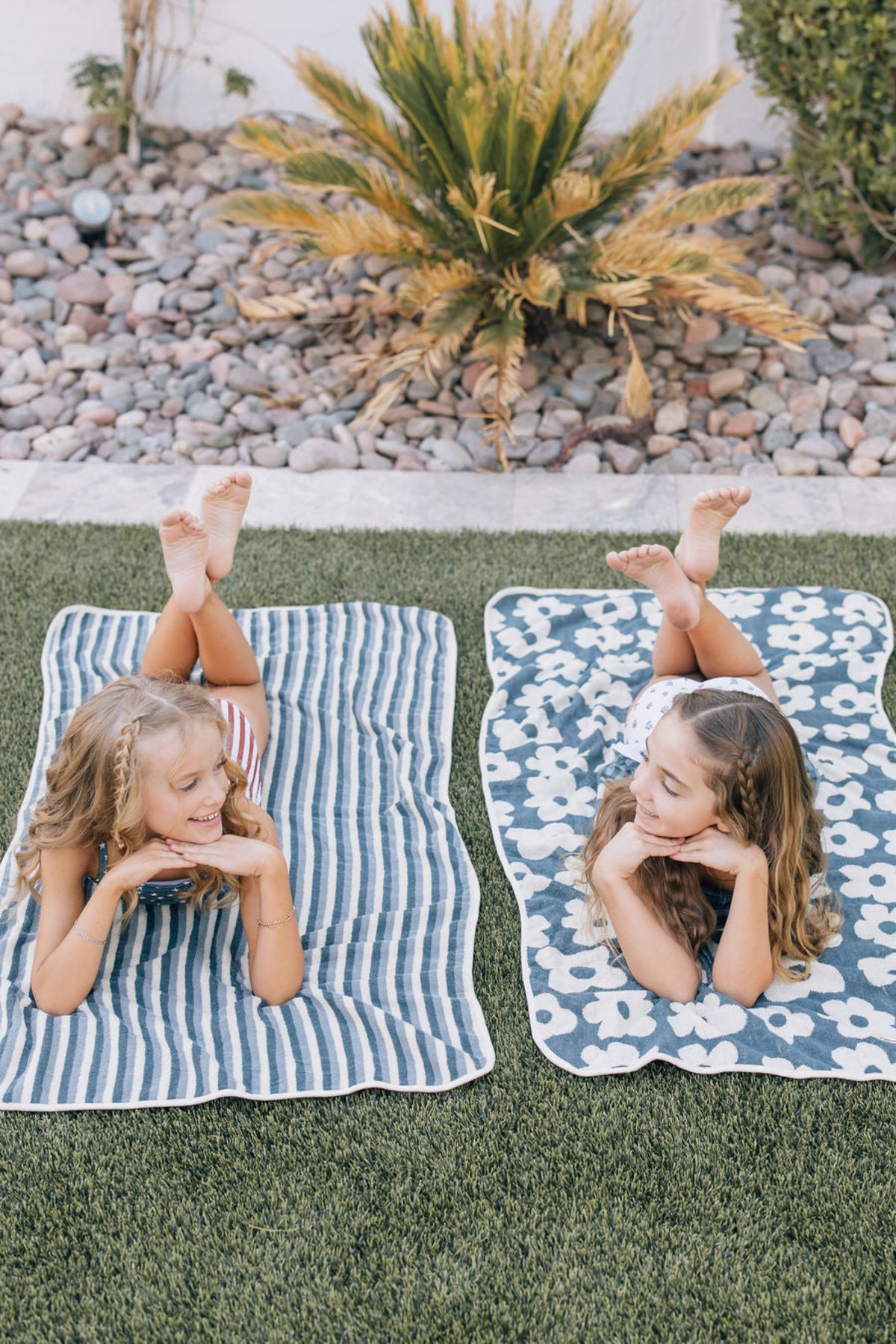 Two young girls lying on navy floral and striped towels on grass, smiling and facing each other playfully.