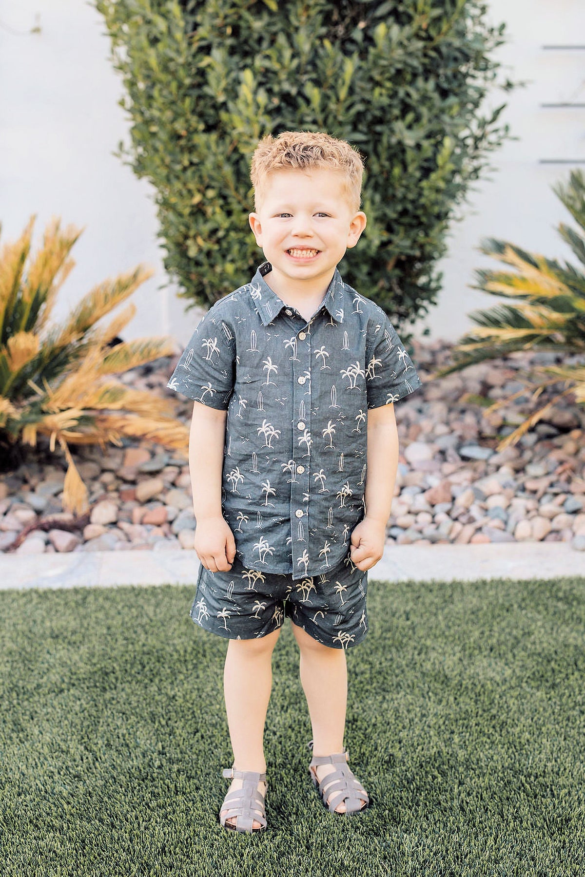 A smiling child wearing a navy shirt and shorts set with palm tree and surfboard print stands on grass.