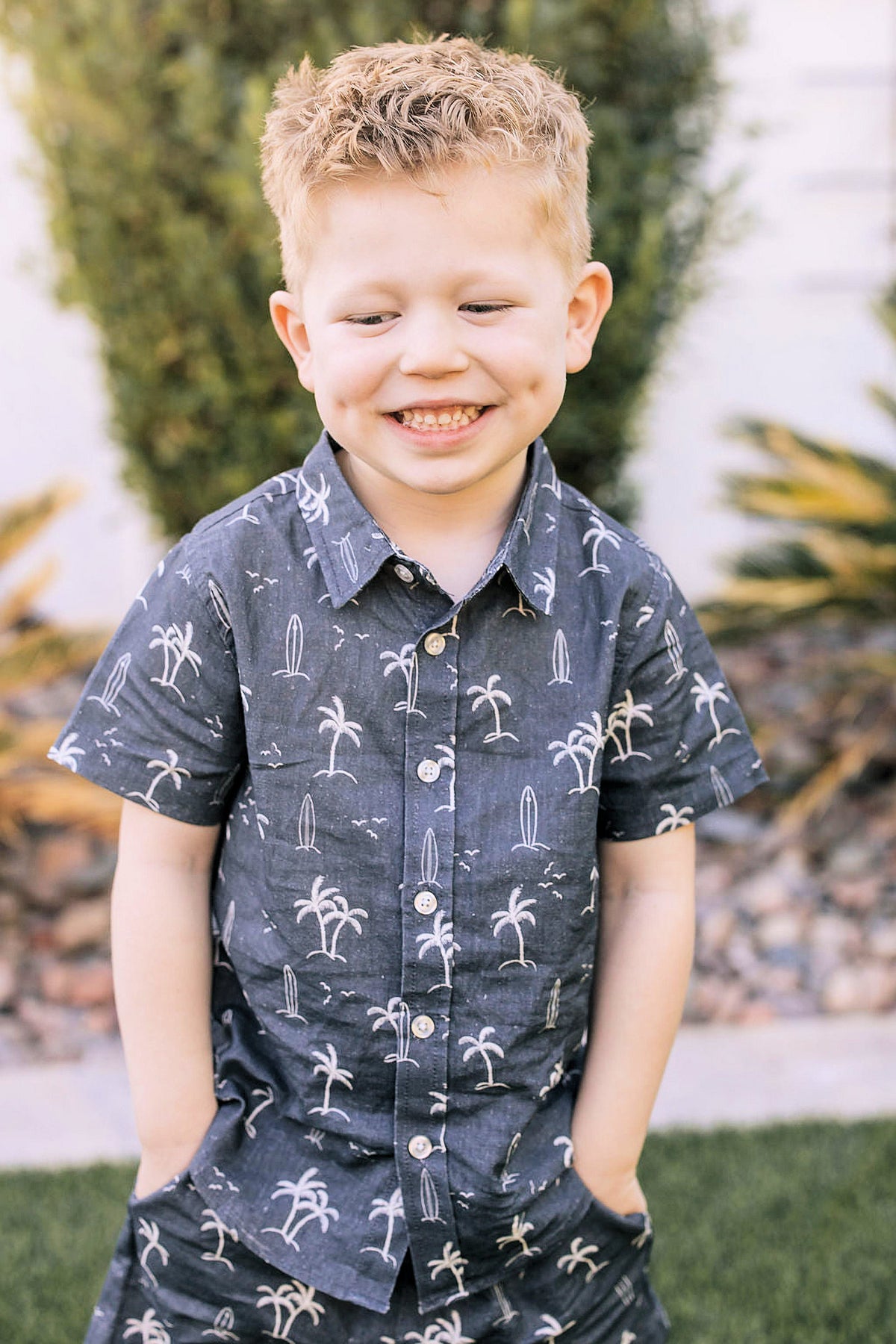 Young boy wearing a navy button-up shirt with surfboard and palm tree print, smiling outdoors in a garden.
