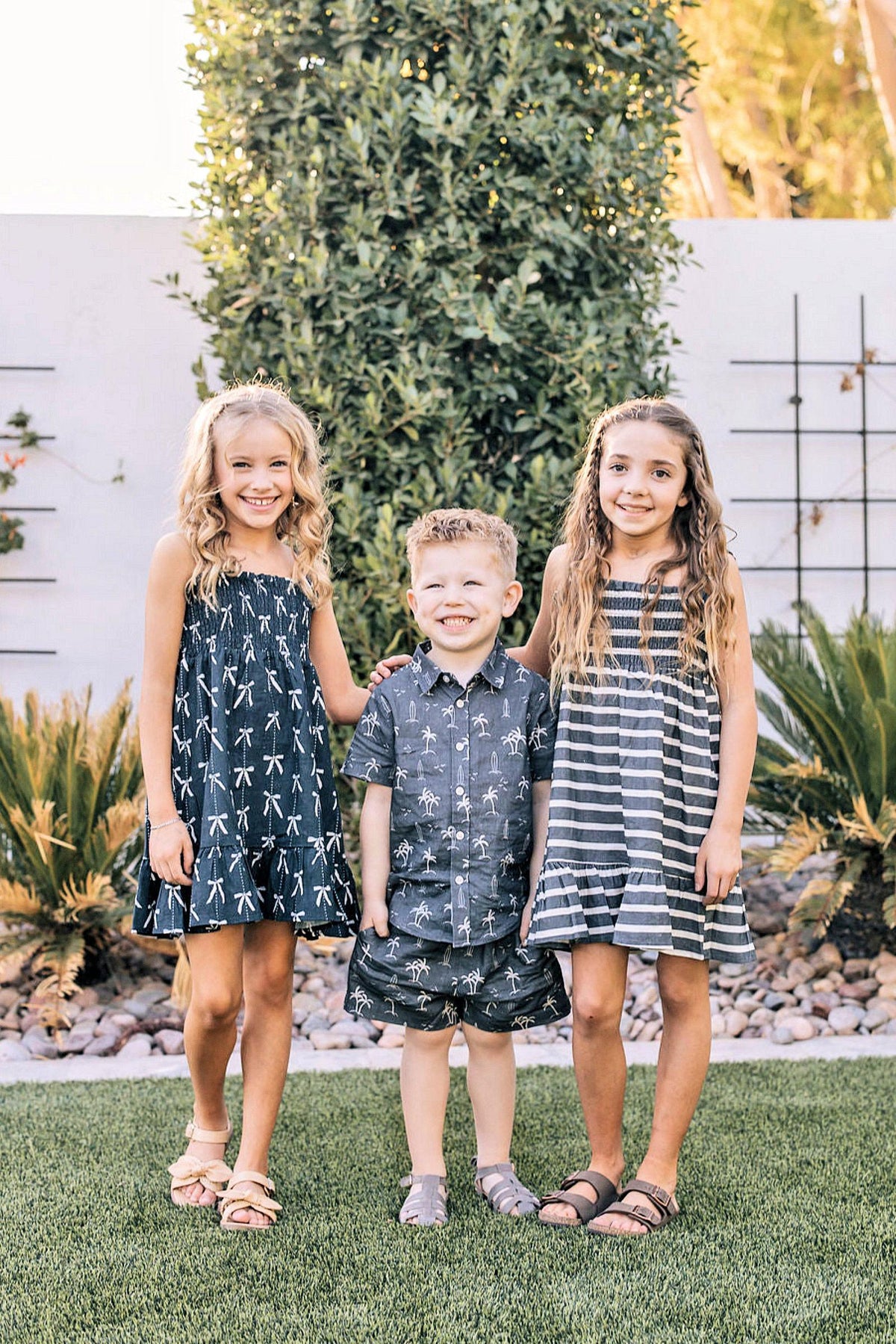 Three children smiling outdoors, wearing summer outfits with patterns, standing on grass with greenery in the background.