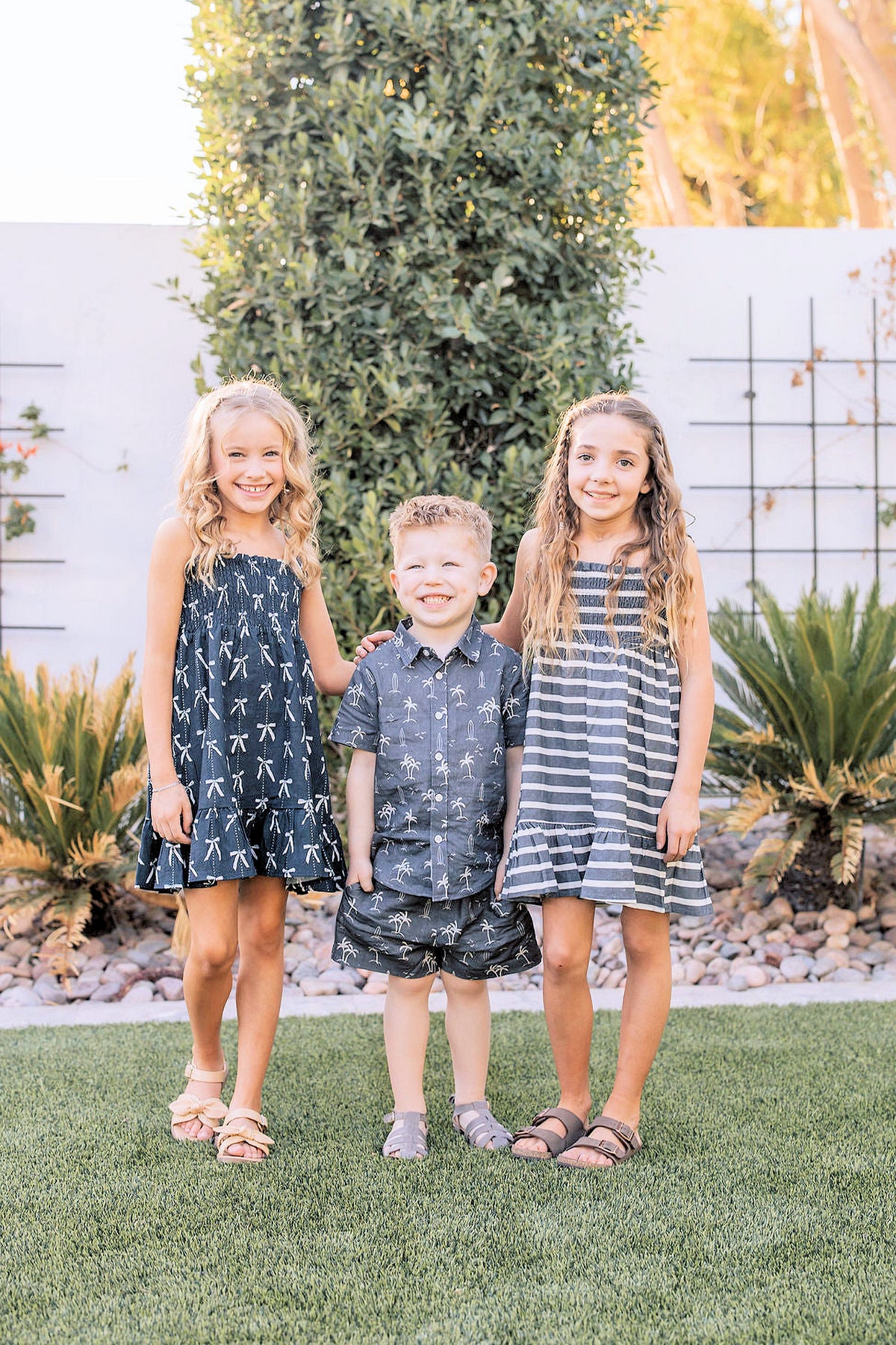 Three children stand on grass wearing navy and striped summer outfits, with greenery and a white wall behind them.