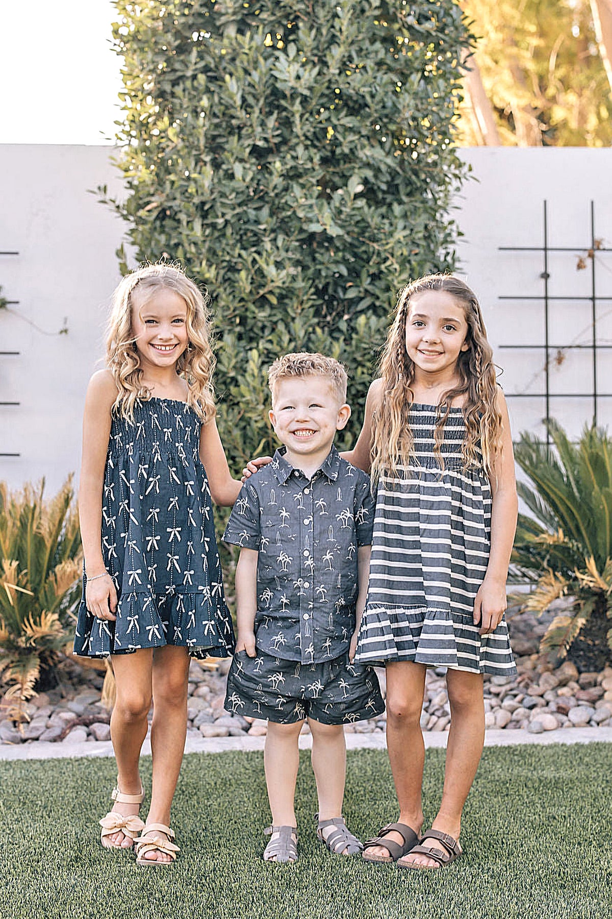 Three children stand on grass, wearing matching navy and striped outfits, with greenery and a white wall in the background.