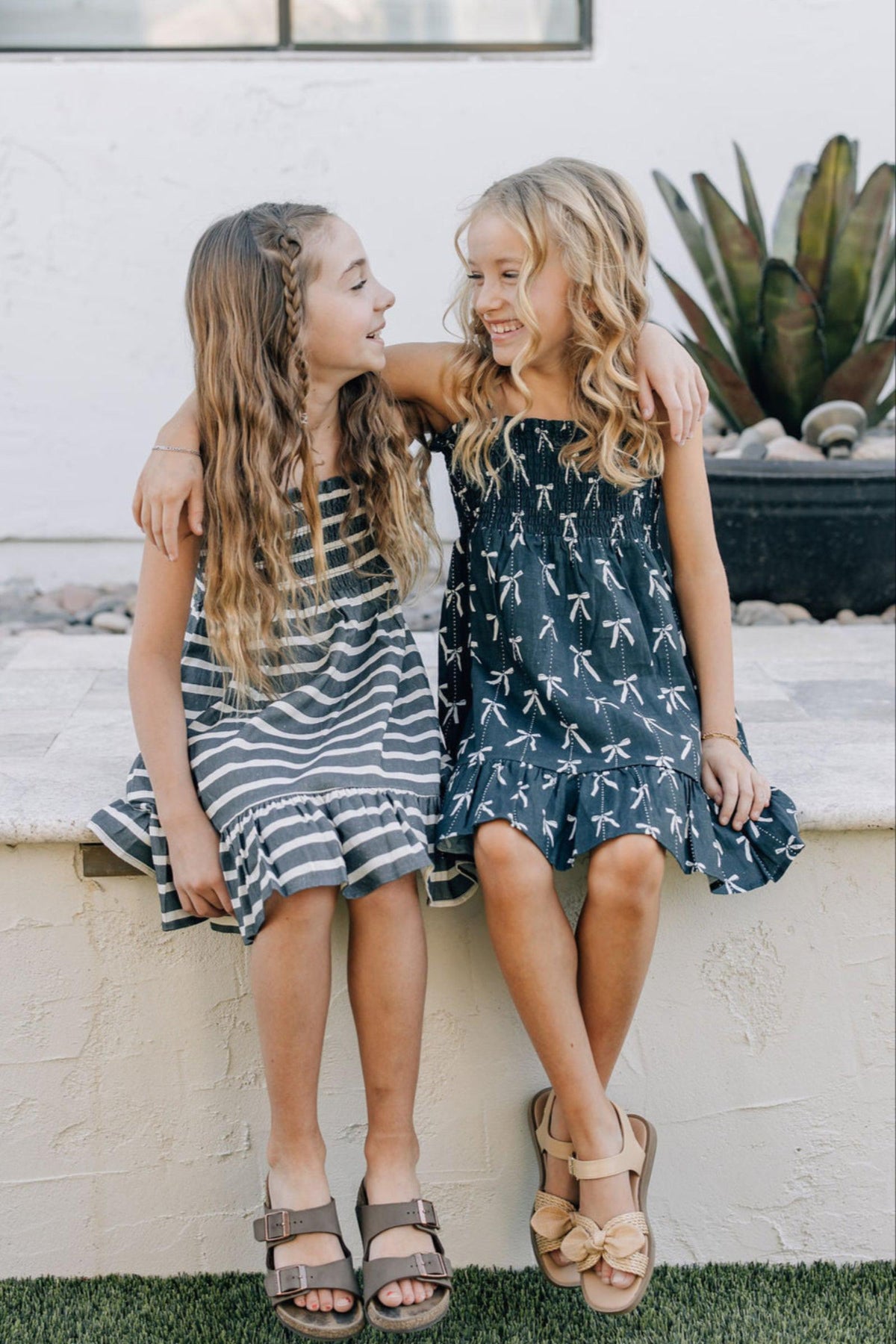 Two girls sitting outside, wearing navy and striped summer dresses, smiling with arms around each other.