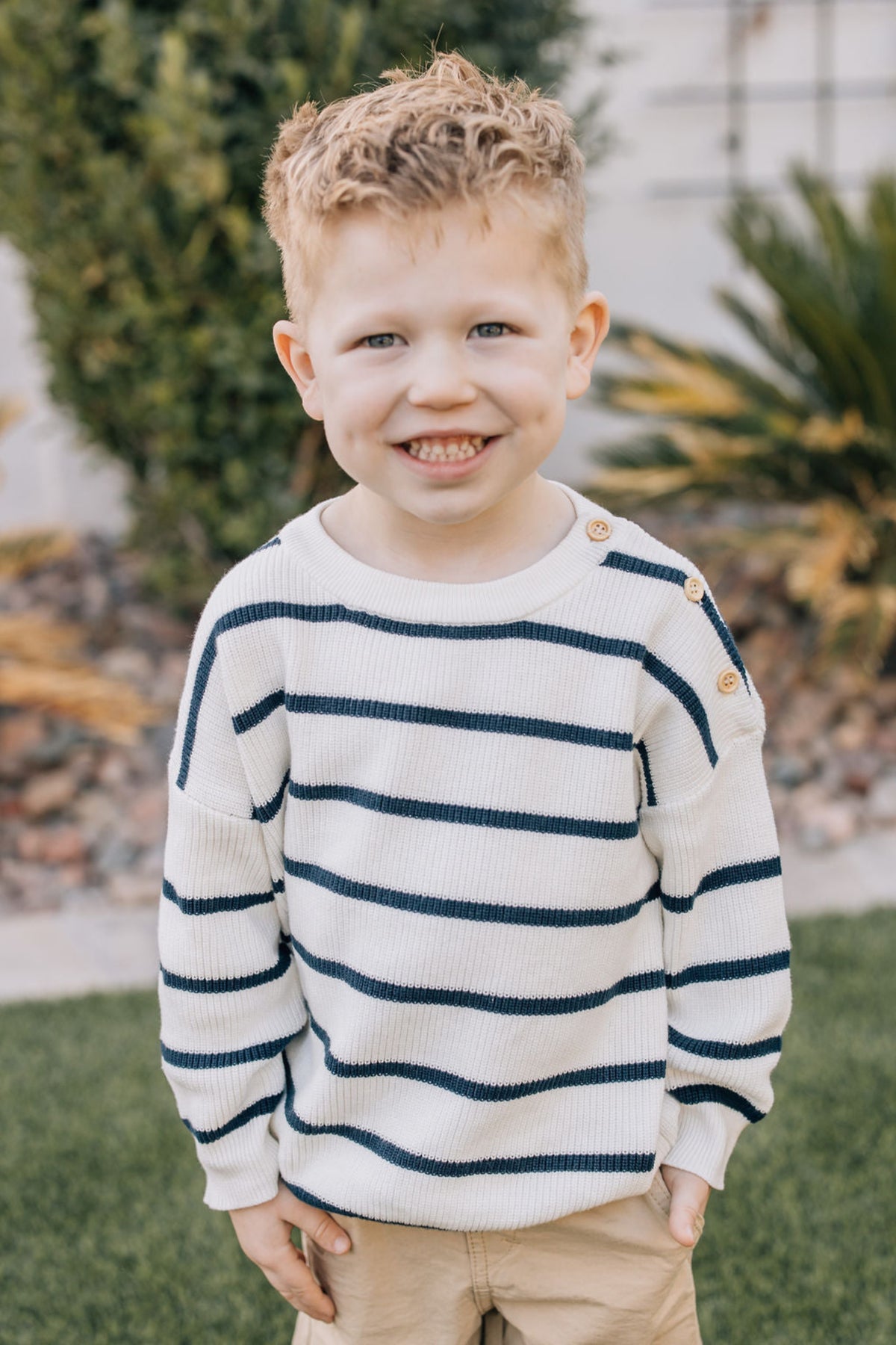 A smiling child outdoors wearing a navy striped knit sweater with button details, standing on grass with plants behind.