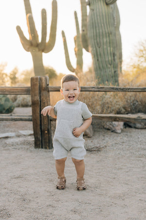 A smiling toddler in grey knit overalls stands in a desert setting with cacti and a wooden fence.