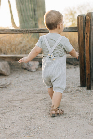 A baby wearing heather grey knit overalls and a grey shirt walks outdoors near a wooden fence.