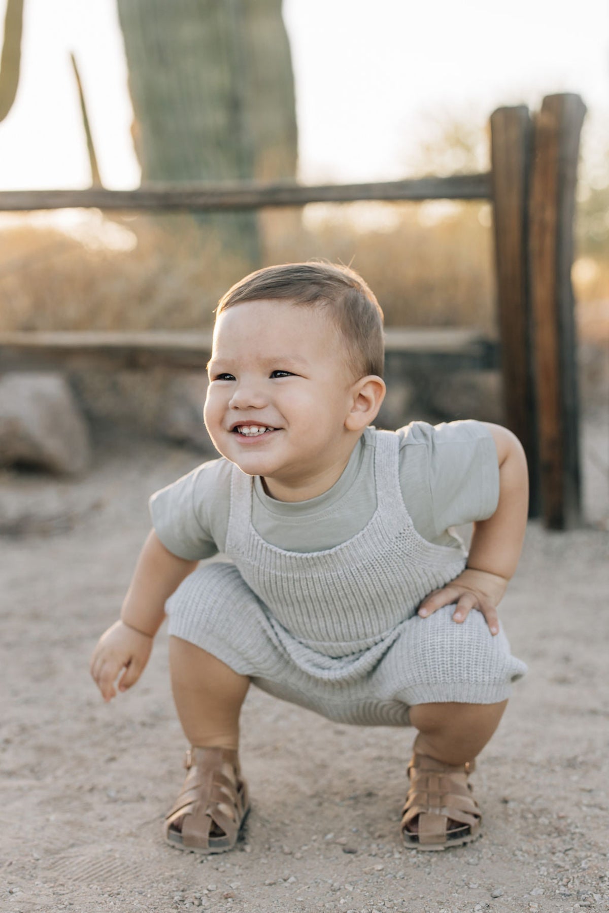 A smiling baby in grey knit overalls crouches outdoors, wearing brown sandals, with a wooden fence behind.