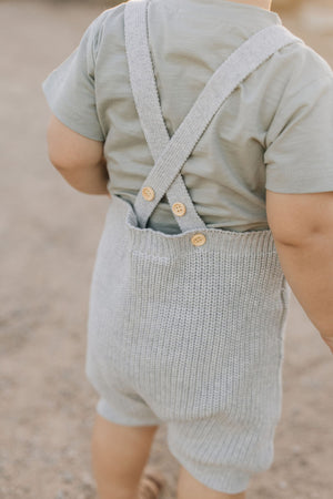 A baby wearing heather grey knit overalls with cross-back straps and wooden buttons, standing outdoors on a sandy surface.