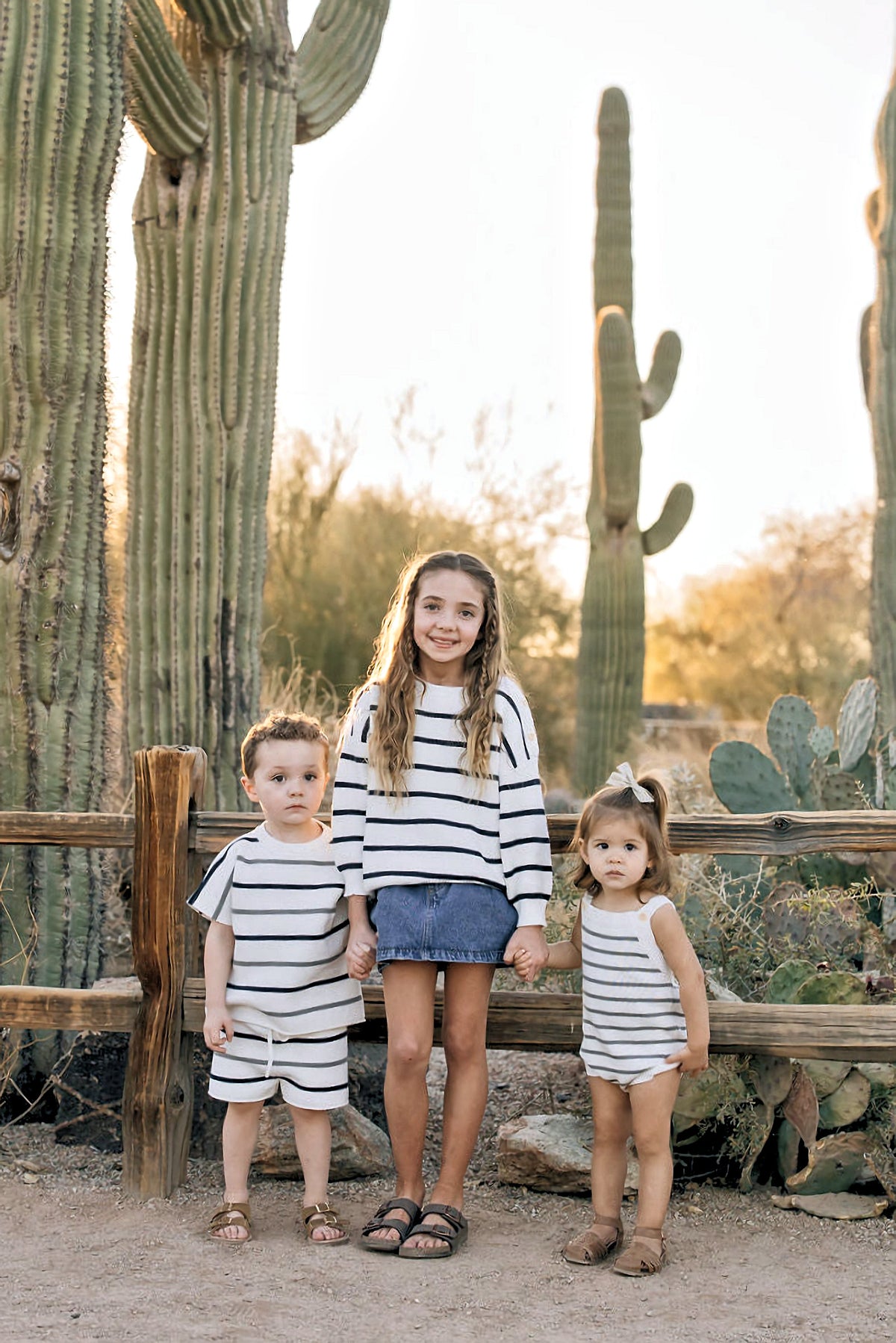 Three children wearing navy stripe knit outfits stand in front of cacti, holding hands and smiling.