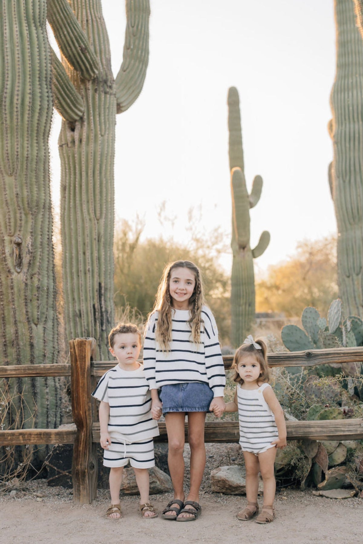 Three children in striped outfits stand together in a desert setting with large cacti and wooden fencing.