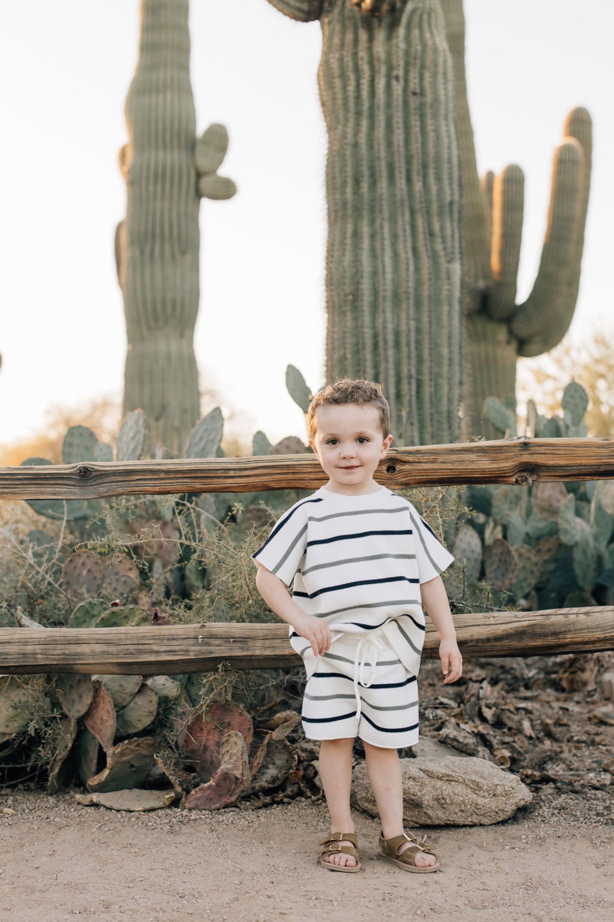 A child wearing a striped knit outfit stands in front of cacti and a wooden fence in a desert setting.