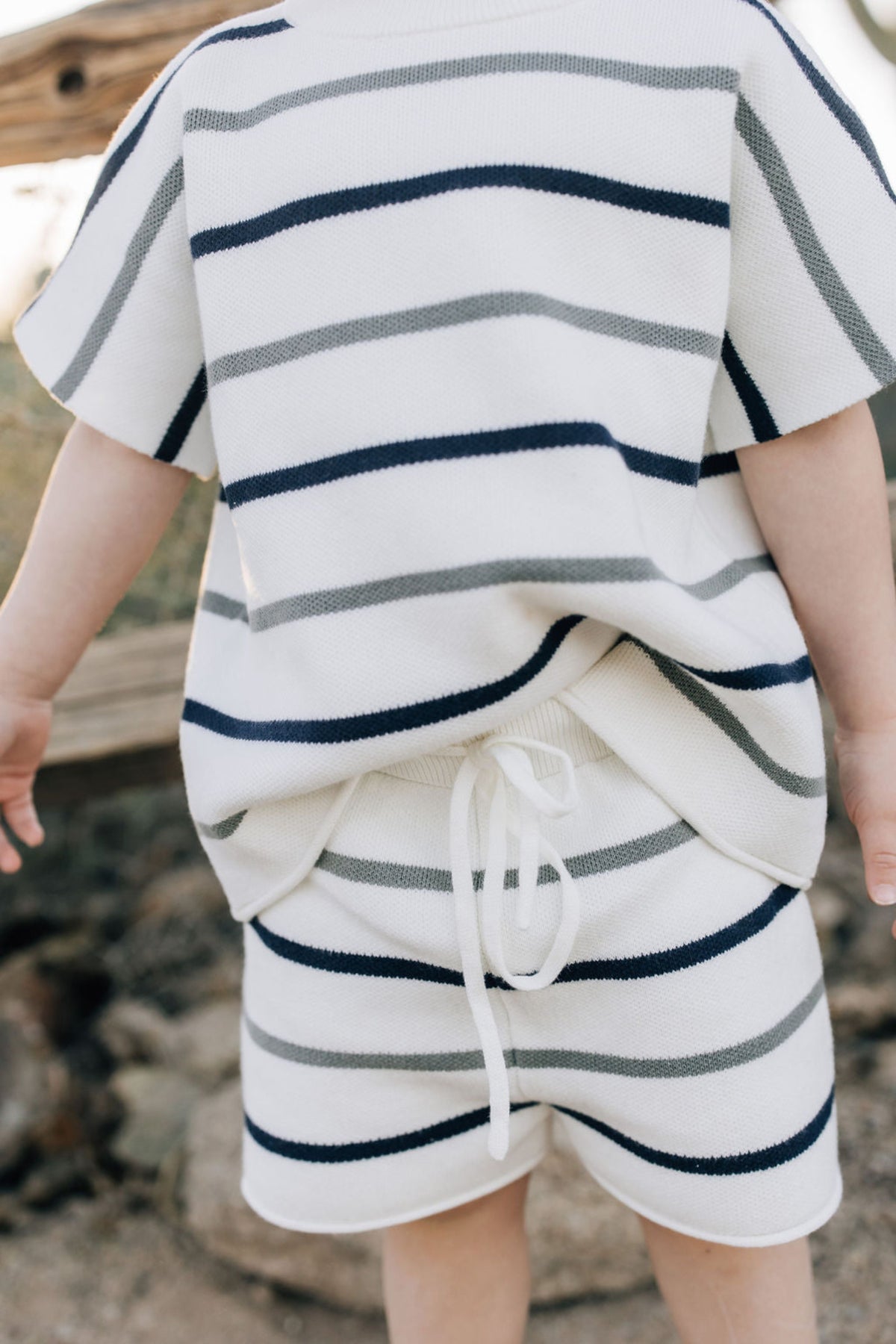 A child wearing a white knit outfit with navy and sage stripes, featuring drawstring shorts outdoors.