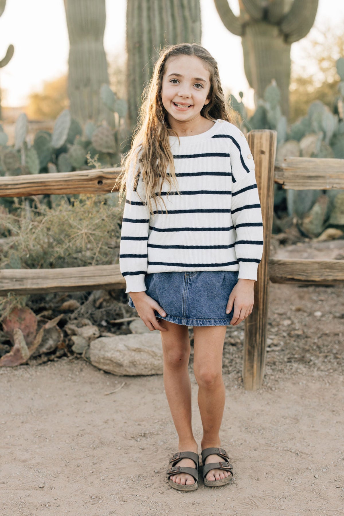 A child wears a navy stripe knit sweater and denim skirt, standing outdoors near cacti with a wooden fence.