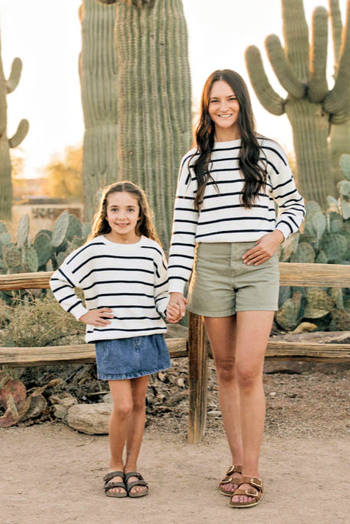 A woman and a child wearing matching navy stripe sweaters stand outdoors in front of tall cacti, smiling.