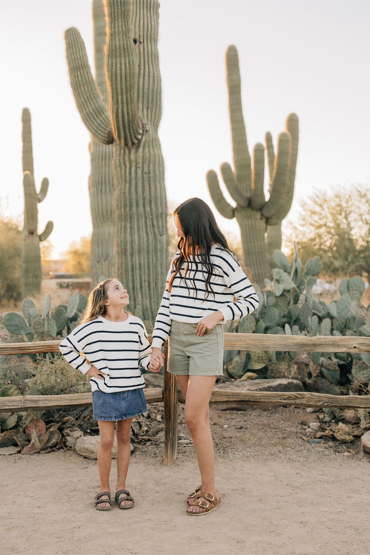 Two people wearing matching navy stripe sweaters stand in a desert landscape with large cacti in the background.
