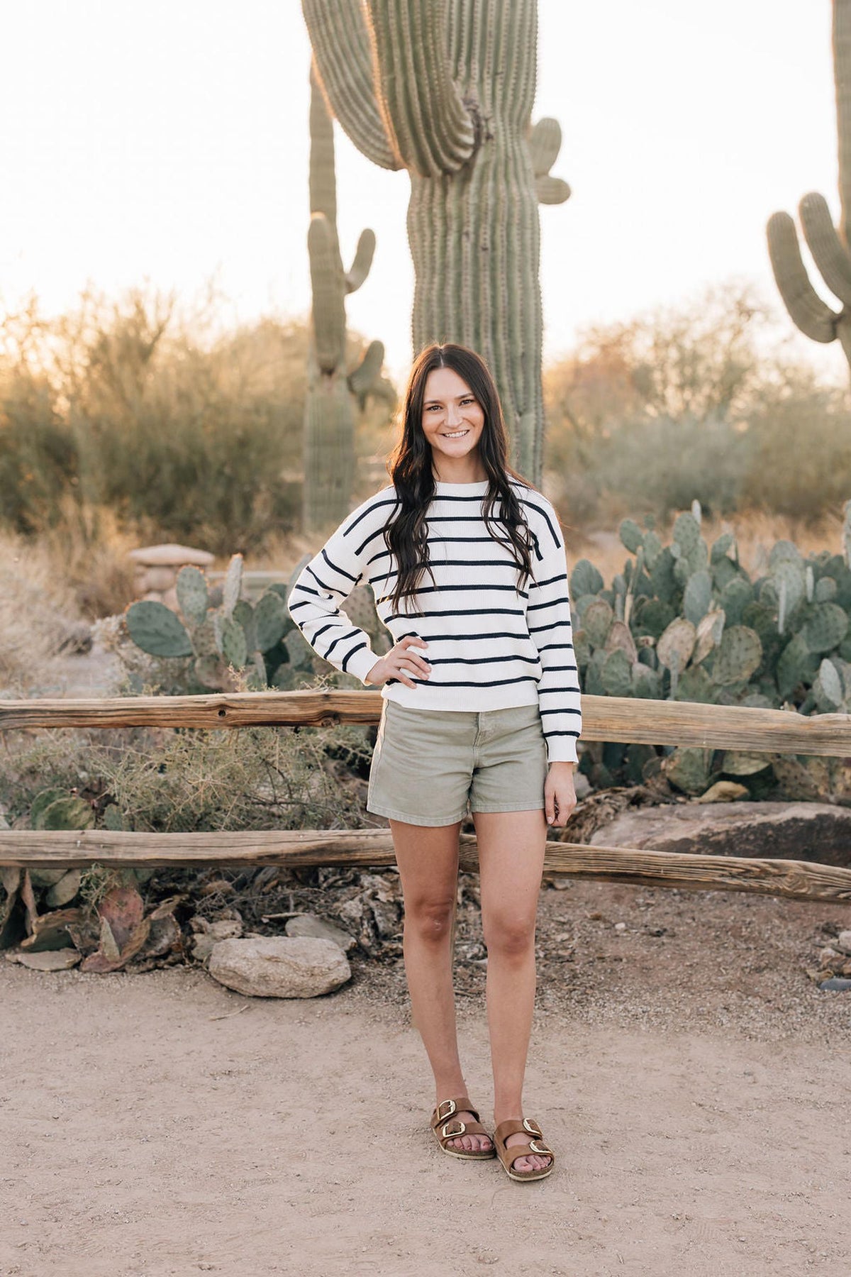 A woman in a navy and white striped sweater stands in a desert landscape with cacti and a wooden fence.