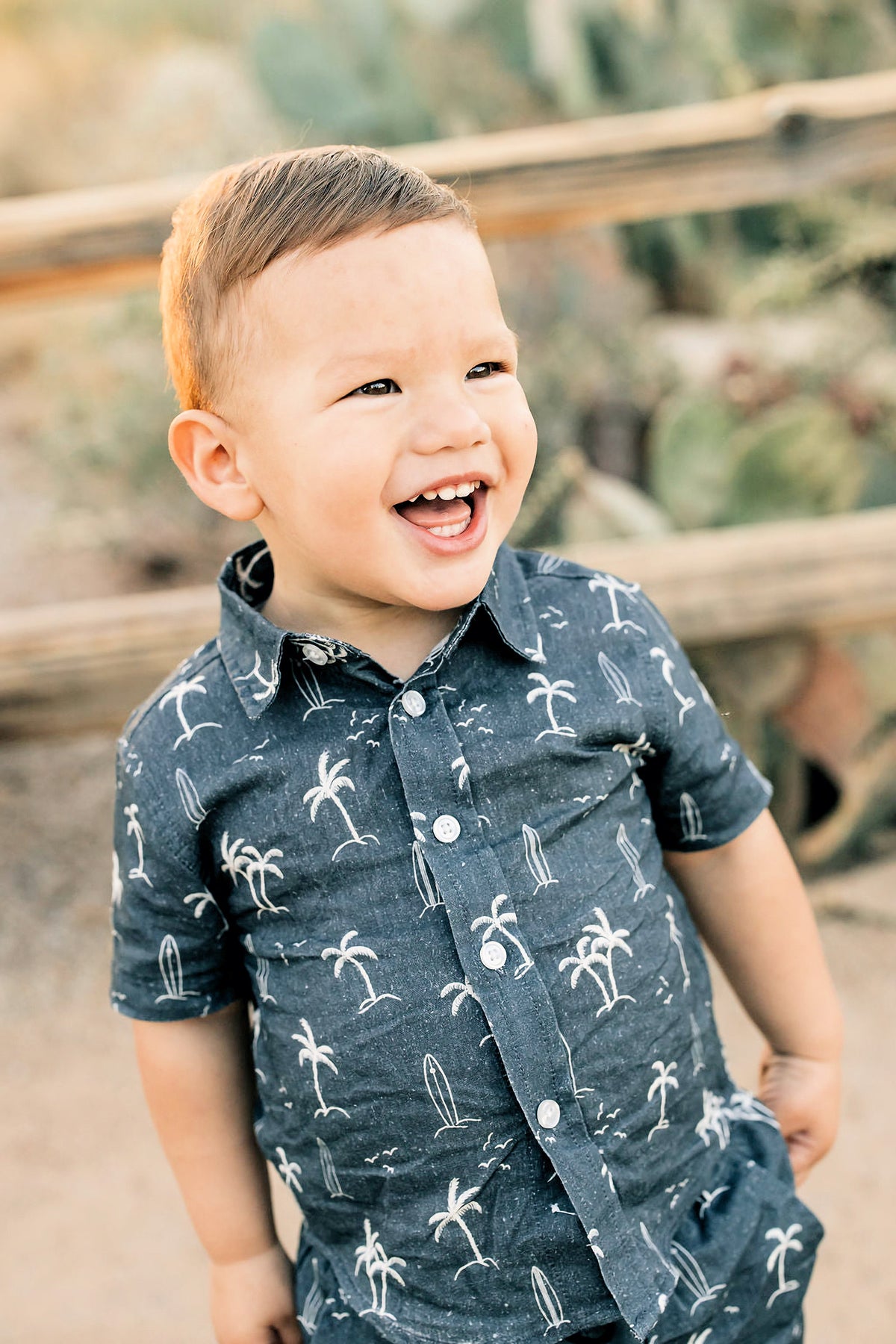 A smiling child wearing a navy button-up shirt with white palm tree and surfboard prints stands outside.