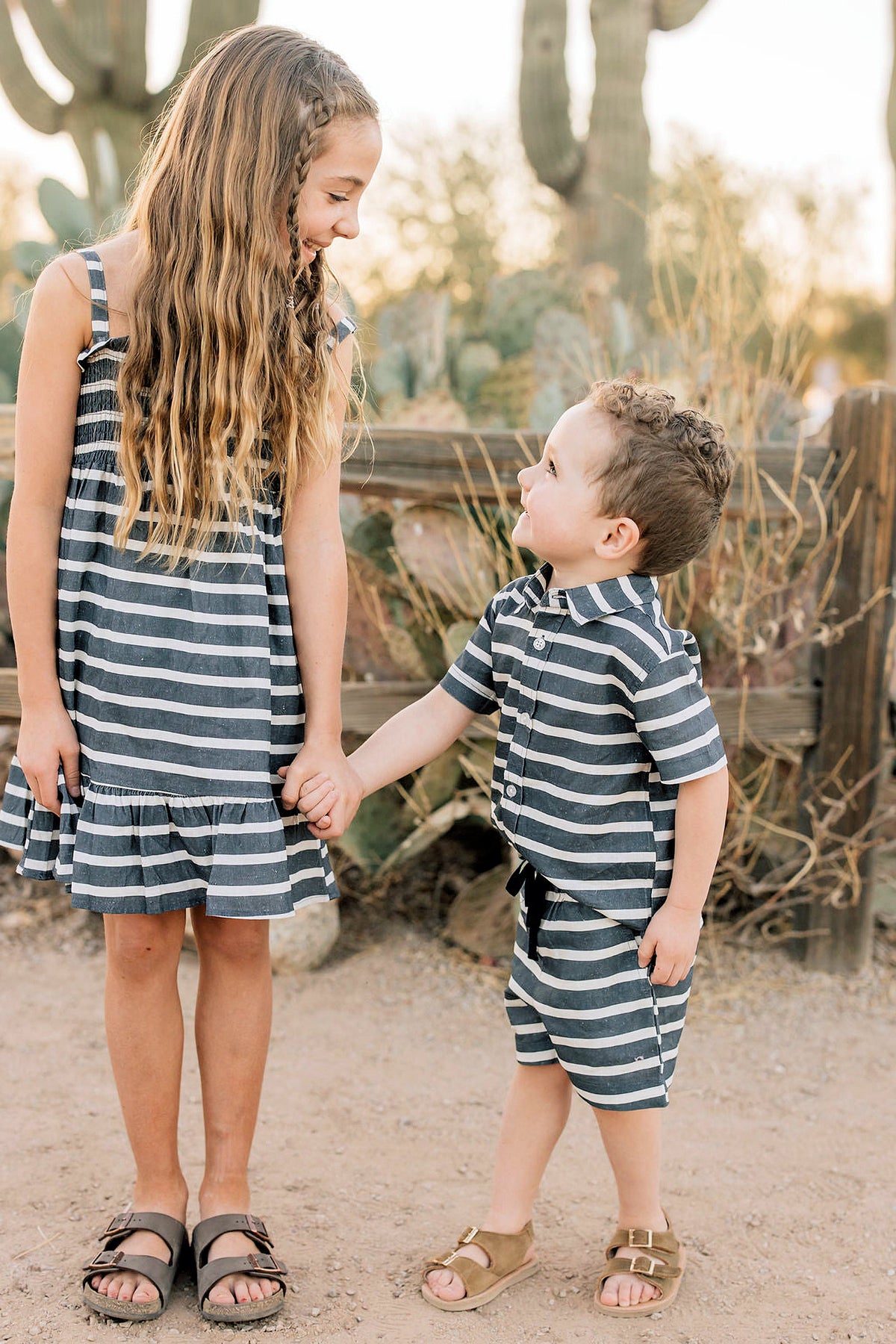 Two children in matching navy and white striped outfits hold hands outdoors, surrounded by cacti and wooden fencing.
