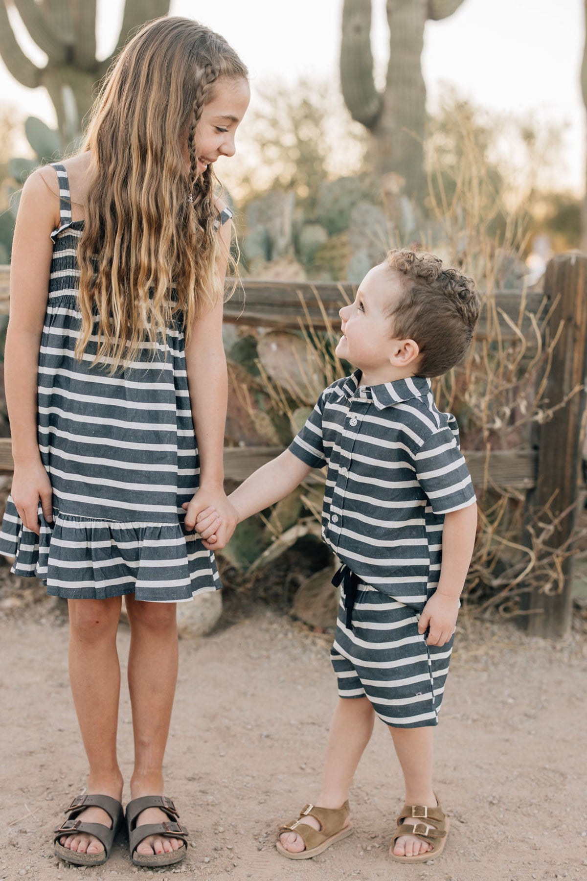 Two children wearing navy and white striped outfits hold hands in a desert setting with cacti.