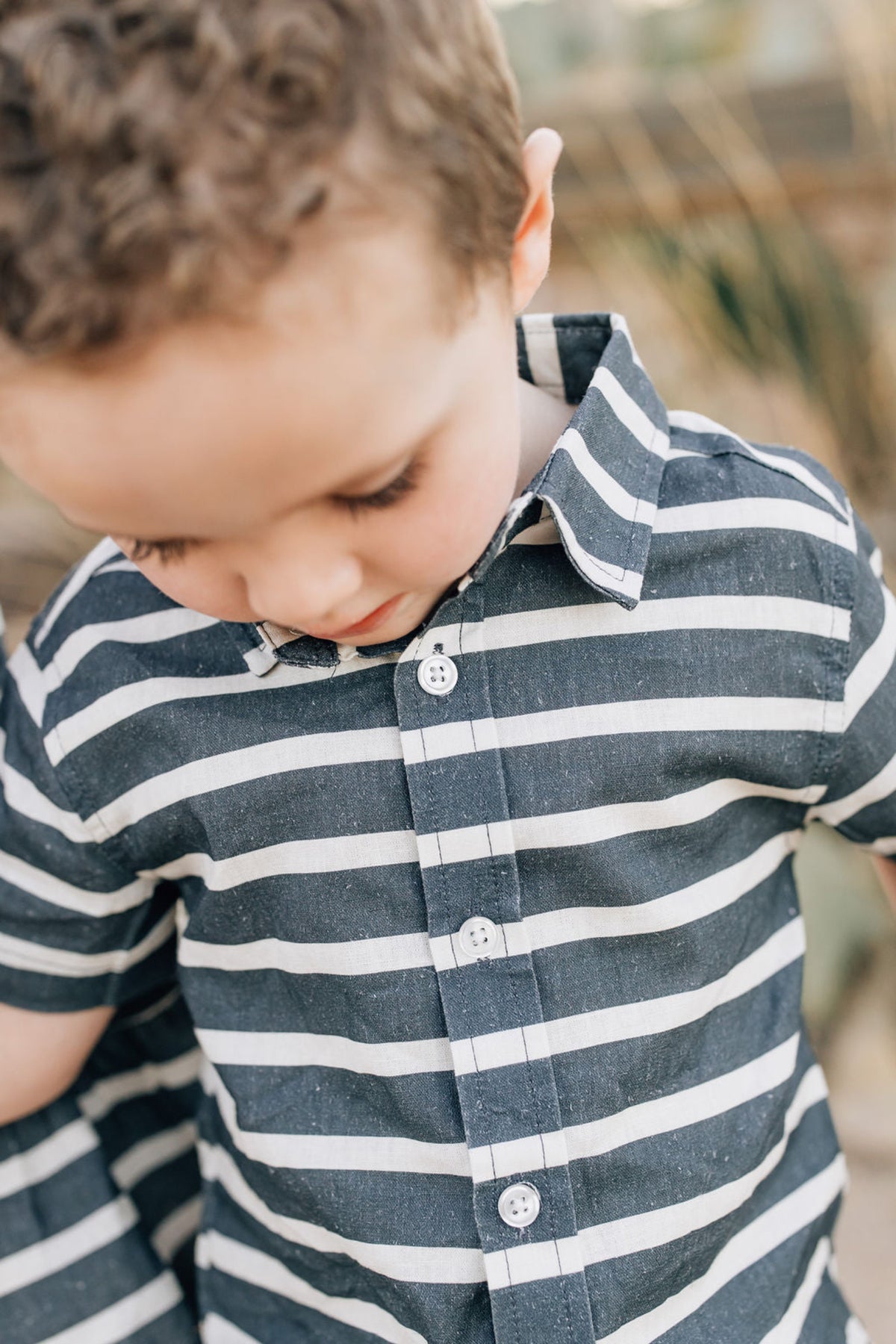 A young child wears a navy and white striped button-up shirt made of linen, looking downwards.
