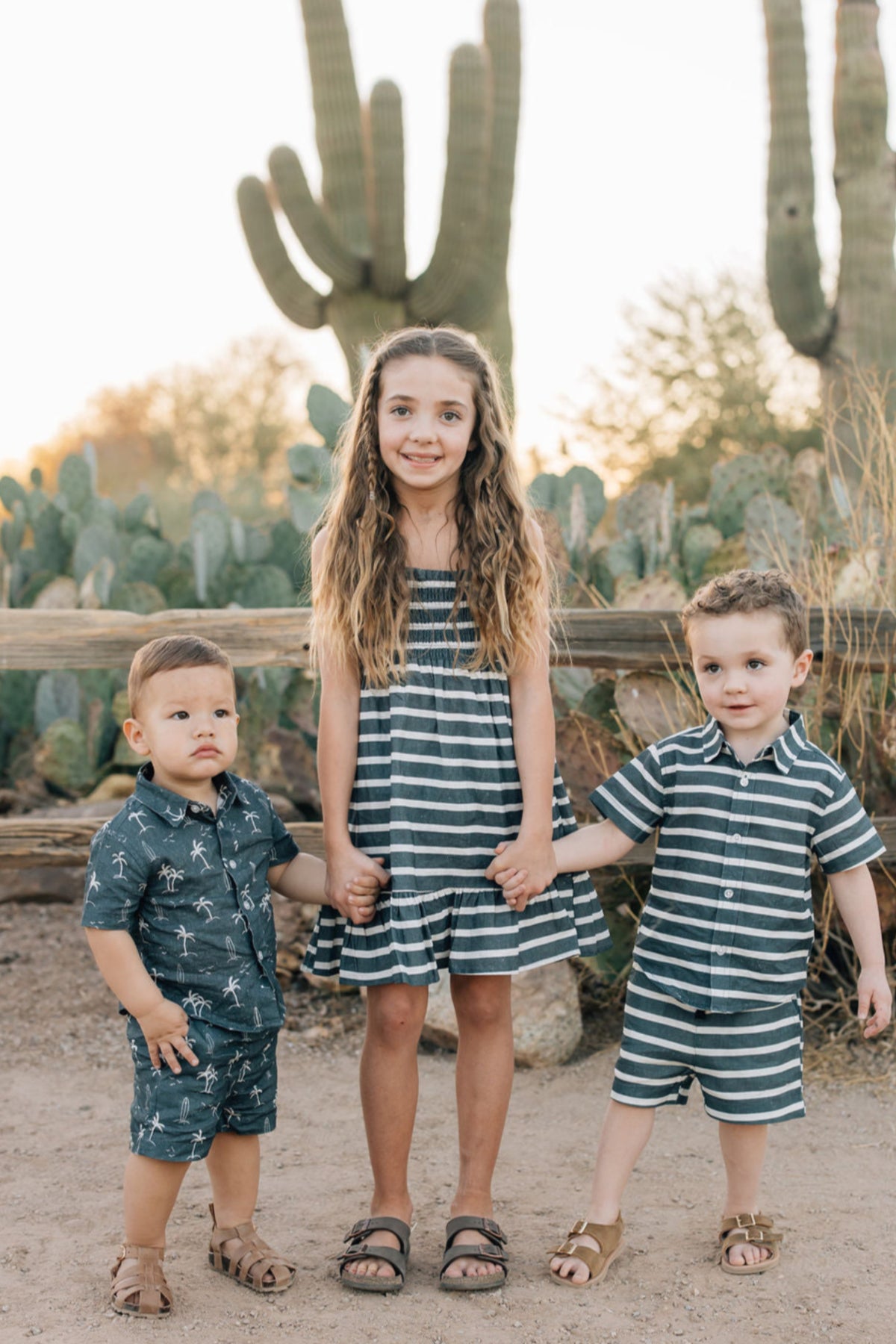 Three children wearing navy and white striped outfits stand in front of cacti, holding hands and smiling.