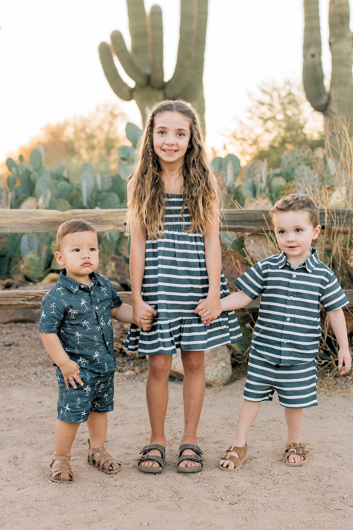 Three children in matching striped and palm print outfits stand in front of cacti, holding hands and smiling.